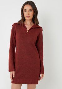 Vrouw in een roestkleurige gebreide jurk met halve rits kraag, staand tegen een effen witte achtergrond.