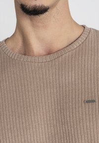 Béžový žebrovaný pletený svetr s kulatým výstřihem a malou kovovou značkou na levé straně. Textura je měkká a mírně elastická.