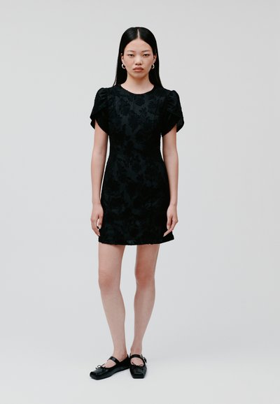 Black dress featuring floral texture, short puff sleeves, fitted silhouette, and above-the-knee length. Worn with black flat shoes.