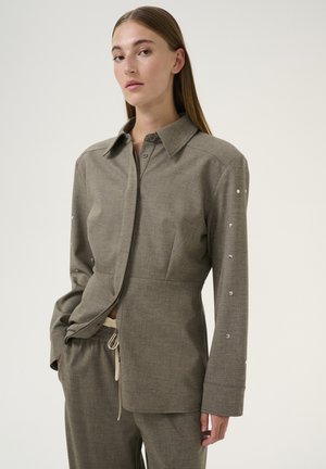 Camicia grigia a maniche lunghe con chiusura a bottone, realizzata in un tessuto strutturato, con borchie decorative lungo le maniche e colletto.
