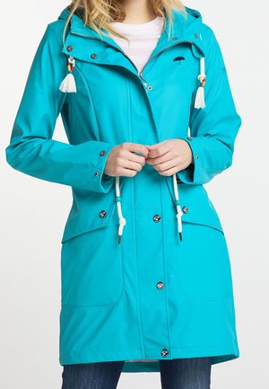 Femme portant un imperméable à capuche bleu avec des cordons blancs, des boutons-pression noirs et des poches kangourou, par-dessus une chemise blanche et un jean.