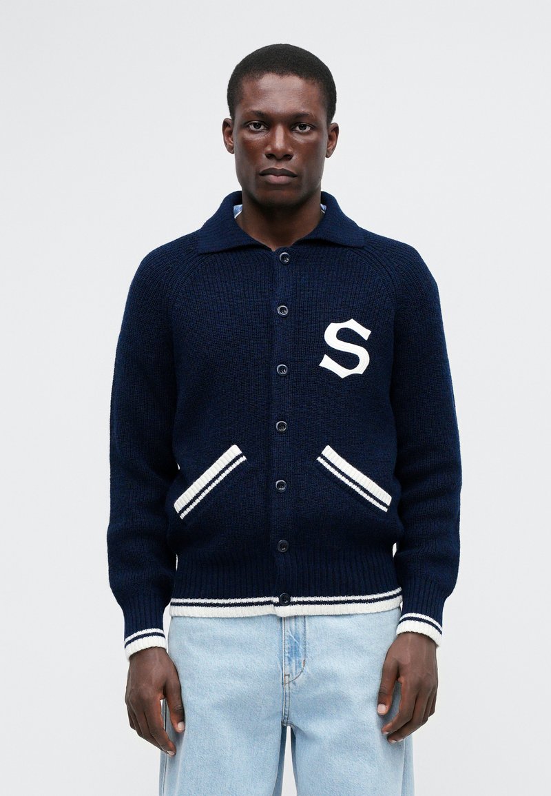 Marineblauwe gebreide cardigan met een kraagnek, zeven knopen, witte accenten op de zakken en manchetten, met een wit 'S' embleem op de borst.