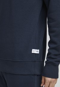 Sudadera azul marino hecha de tela suave, con puños y dobladillo acanalados, y una pequeña etiqueta de marca blanca en la parte inferior izquierda.