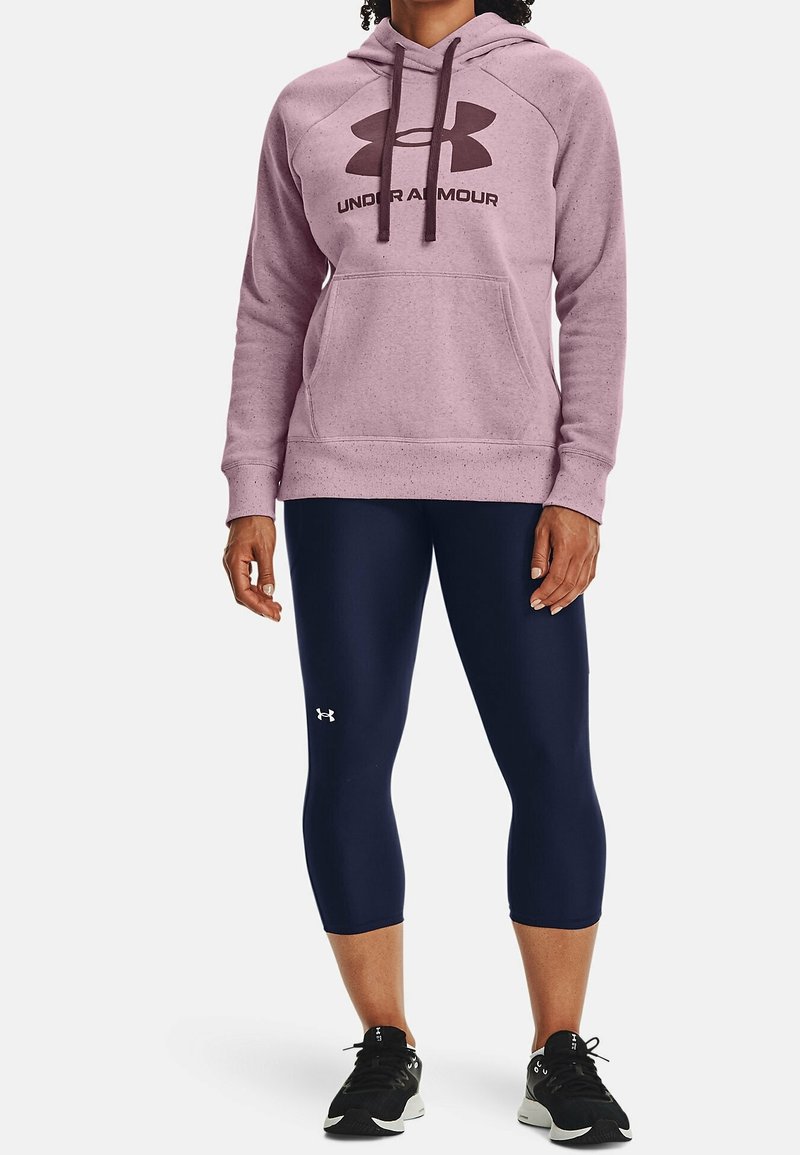 Personne portant un sweat à capuche rose Under Armour, des leggings courts bleu marine avec logo et des chaussures de sport noires, debout devant un fond blanc.