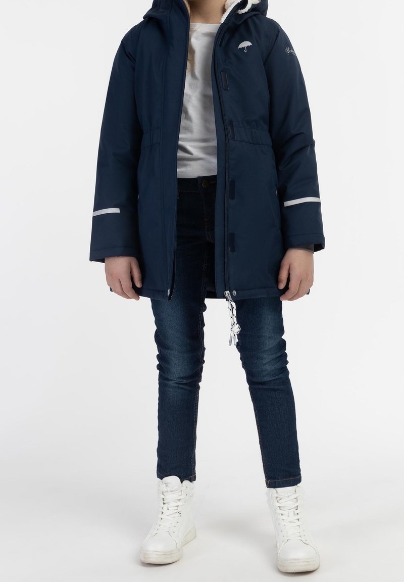 Enfant portant une parka bleu marine avec une capuche bordée de fourrure, un jean bleu foncé, des baskets blanches et une chemise blanche, debout devant un fond blanc.