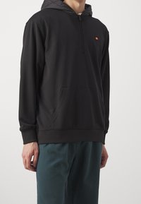 Felpa nera con zip, realizzata in tessuto leggero, dotata di tasca a marsupio e di un piccolo logo sul petto. Abbinata a pantaloni color teal.