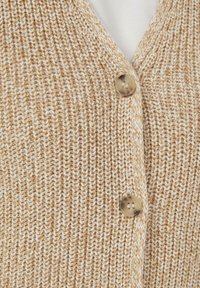 Beige en witte gebreide cardigan met een verticale ribbelpatroon en twee lichtbruine knopen op een witte onderguling.