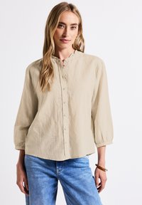 Ljusbeige skjorta med knappar, rund krage, trekvartsärmar och strukturerad tyg. Kombinerad med blå jeans.