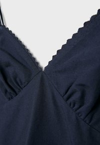Tissu bleu marine avec encolure bordée en forme de festons et coutures froncées sur un vêtement sans manches, sur un fond clair uni.