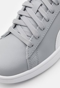 Šedá kožená teniska s hladkou texturou, plochými šedými tkaničkami a bílou gumovou podrážkou, s prošitými detaily po straně.