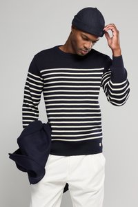 Navy-blau gestreifter Pullover mit cremfarbenen horizontalen Streifen, runder Ausschnitt und Knopfdetails an der Schulter, kombiniert mit weißen Hosen.