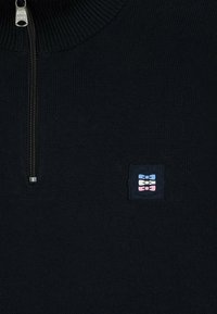 Pull en tricot bleu marine avec fermeture zippée à mi-hauteur, col côtelé et logo en patch rectangulaire sur le côté gauche. La texture est rugueuse et épaisse.