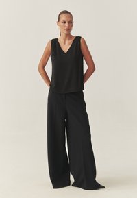 Črni sleeveless top z V-izrezom in dekorativno teksturo, v kombinaciji z visokim pasom črnih širokih hlač s sproščenim pristopom.