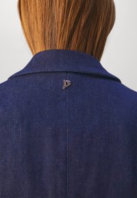 Cheveux lisses châtain clair arrivant aux épaules, dépassant le col d'une veste en jean bleu foncé avec une petite épingle argentée en forme de "D" au centre du dos.