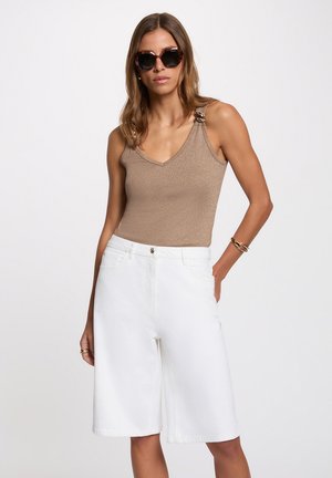 Femme portant de grandes lunettes de soleil foncées, un top beige sans manches avec des bretelles décoratives, un short blanc taille haute longueur genou et un bracelet doré.