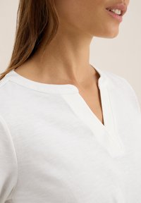 Femme portant un t-shirt blanc en V, mettant en valeur le cou, la mâchoire et une partie de ses longs cheveux bruns, sur un fond neutre.