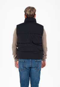 Zwarte gewatteerde vest met hoge kraag, segmentontwerp, gedragen over een lichtbruine lange mouw shirt, gecombineerd met blauwe jeans.