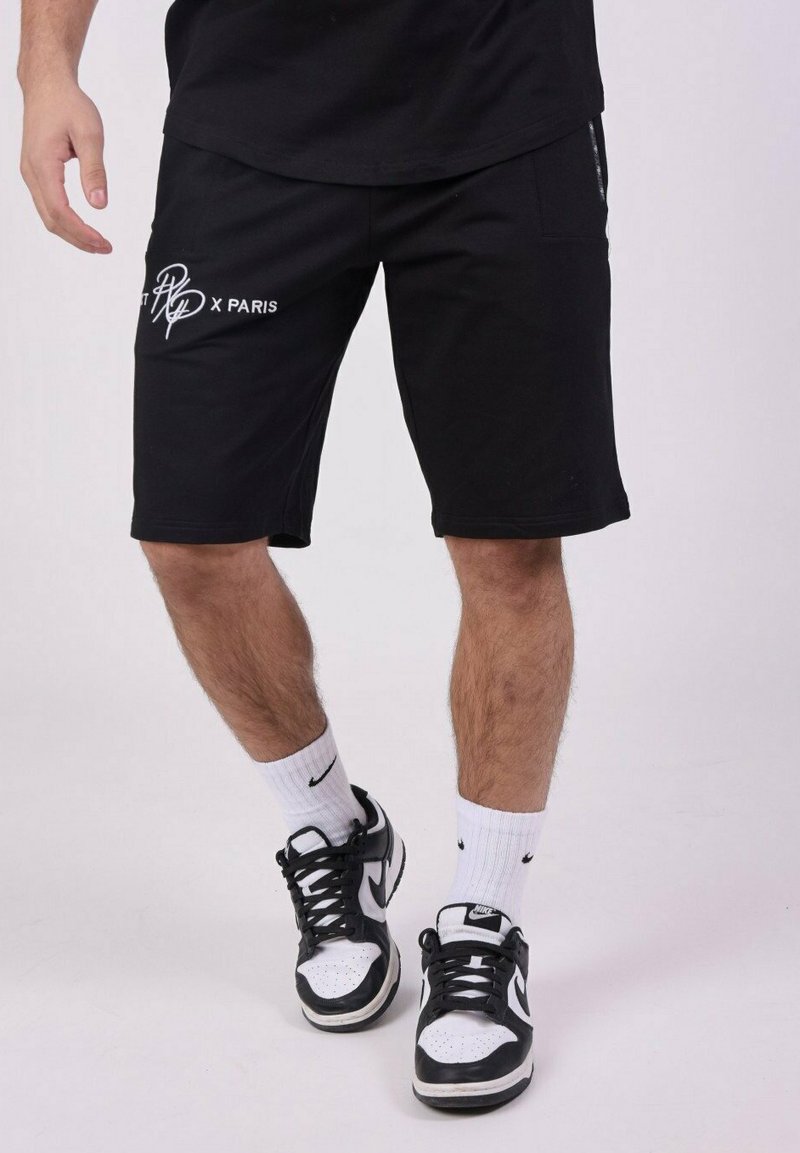 Pantalones cortos deportivos negros con un corte relajado, que cuentan con bolsillos laterales y un texto gráfico blanco "PT x PARIS". Combinados con zapatillas negras y blancas.