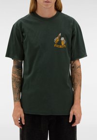 Osoba w ciemnozielonym t-shircie z grafiką przedstawiającą szkielety trzymające butelkę piwa i kufel nad napisem "VANS 1966" na klatce piersiowej.
