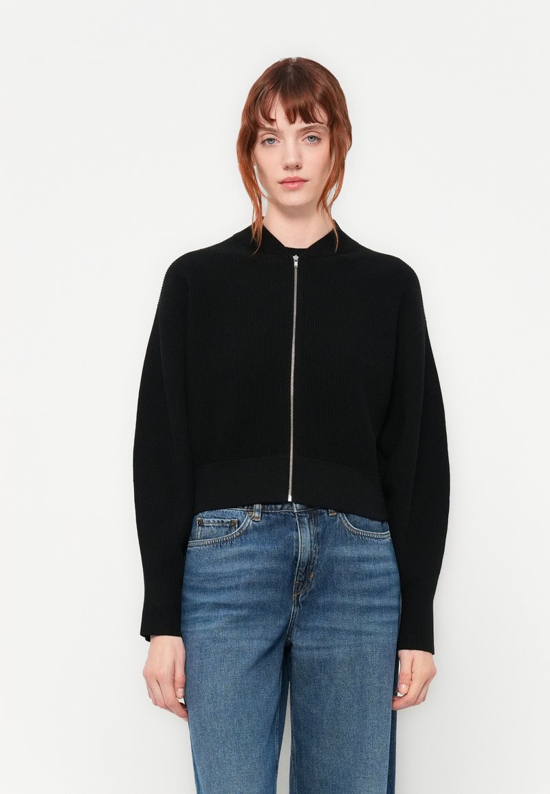 Svart ribbestrikket genser med en kort design, høyt krage og detaljer med sølvglidelås, kombinert med lyseblå rette jeans.