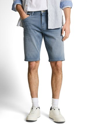 Man met blauwe spijkershorts, witte sneakers met witte sokken, een wit T-shirt en een opgerold lichtblauw overhemd met knopen.