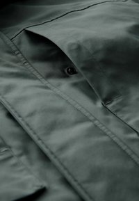 Tejido de chaqueta verde oscuro, con una textura suave, costuras visibles, un bolsillo con tapa y un detalle de botón negro.