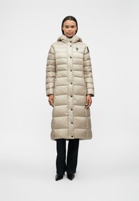 Lunga giacca trapuntata beige con cappuccio, caratterizzata da cuciture orizzontali, bottoni frontali e una toppa sulla manica. Indossata sopra dei jeans scuri.