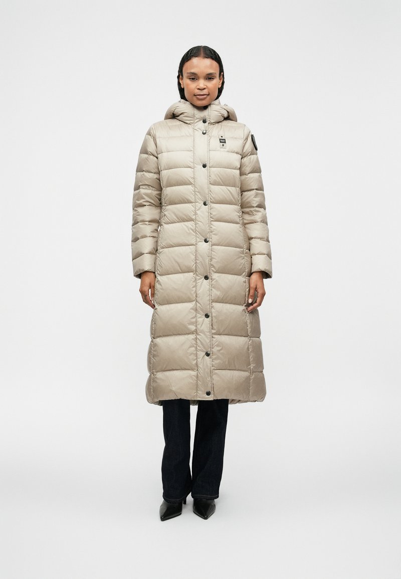 Lunga giacca trapuntata beige con cappuccio, caratterizzata da cuciture orizzontali, bottoni frontali e una toppa sulla manica. Indossata sopra dei jeans scuri.