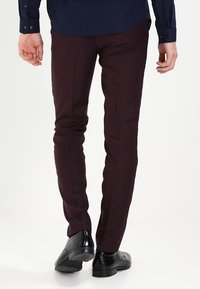 Burgundy slim-fit byxor med en slät yta, med en skräddarsydd design och extra detaljer längs sömmarna. Svarta dress-skor synliga.