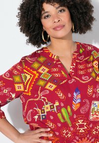 Blusa roja con estampado geométrico y motivos de la naturaleza, que presenta colores vibrantes como verde, amarillo, azul y blanco. Corte holgado y mangas cortas.