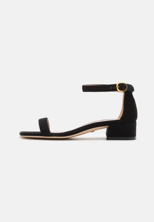 Black suede low block heel sandal with ankle strap and gold buckle, open toe and minimalist design on white background.