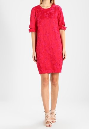 Robe rouge longueur genou avec texture florale, manches retroussées aux coudes, portée avec des talons beiges à brides croisées.
