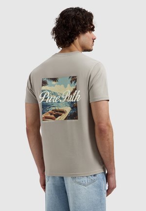Man met krullend haar die een beige T-shirt draagt met een boot- en oceaangrafiek en de tekst "Pure Path" op de achterkant, gecombineerd met lichtblauwe spijkerbroek.