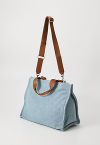 Blå denimtaske med brune læderhank og justerbar rem, der har en struktureret form og blød tekstur. Sølv hardware detaljer inkluderet.