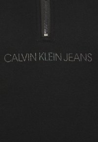 Parte superior negra con cremallera y el logo "CALVIN KLEIN JEANS" en letras iridiscentes. Tela suave con textura lisa. Detalle de cremallera metálica.