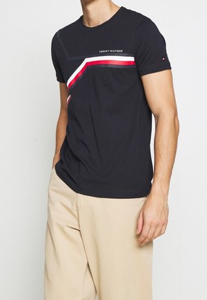 Mężczyzna noszący czarną koszulkę Tommy Hilfiger z czerwonymi, białymi i niebieskimi skośnymi paskami, w połączeniu z beżowymi spodniami, stojący z rozluźnionymi rękami.
