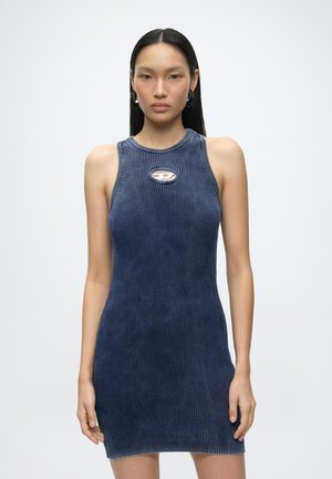 Giovane donna con capelli neri lisci che indossa un abito blu navy aderente senza maniche in maglia a coste, con un piccolo dettaglio a ritaglio sul petto.