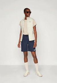 Lengvas, kremas spalvos pūkuotas liemenė virš bežinio marškinėlio, derinama su tamsiai mėlynomis šortais ir bežinėmis sportinėmis bateliais. Akiniai nuo saulės užbaigia sportišką įvaizdį.