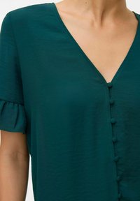 Blusa teal a maniche corte con scollatura a V, caratterizzata da una patta con bottoni rivestiti in tessuto e una ruffle arricciata sulle maniche.