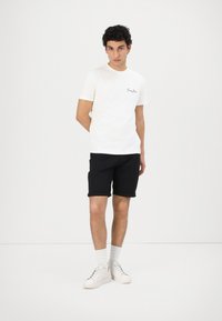 White short-sleeve t-shirt with embroidered logo, paired with black shorts. Model wears white sneakers and ribbed ankle socks. Simple design.