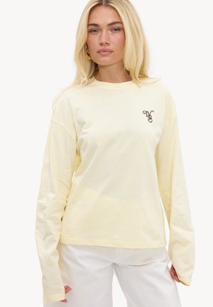 Blondynka w bladoróżowej żółtej koszuli z długim rękawem z małym czarnym monogramem na lewej piersi oraz białych spodniach.