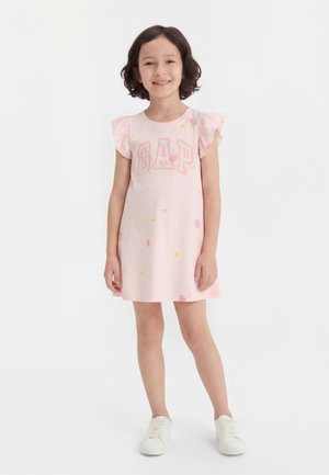 Jong meisje lacht, draagt een lichtroze jurk met geruite mouwen, GAP-logo, kleine fruitpatronen en witte sneakers, staand op een witte achtergrond.