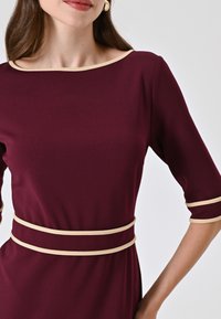 Abito bordeaux con scollatura a barca, maniche a tre quarti e accentuazioni beige lungo il collo e in vita. Realizzato in tessuto liscio.