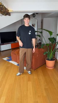 Crna majica s bijelim grafičkim uzorkom, smeđe cargo hlače i srebrne cipele; unutarnji prostor s biljkom u tegli i narančastom sofom.