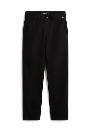 Person iført en hvid Vans T-shirt, sorte bukser, blå Vans sneakers og en lilla sweatshirt bundet omkring livet med en ternet stribe.