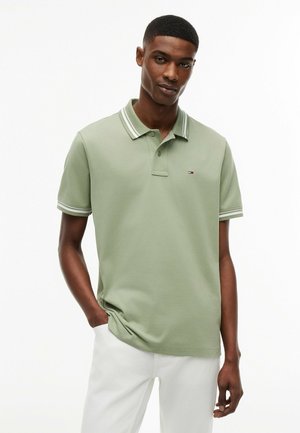 Junger Mann mit kurzen Haaren, der ein hellgrünes Poloshirt mit weißen Streifen am Kragen und an den Ärmeln trägt, kombiniert mit weißen Hosen, steht entspannt.