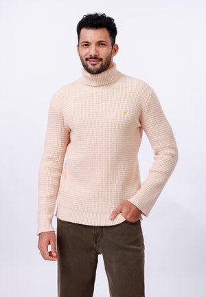 Suéter de punto waffle con cuello alto en rosa suave, con un dobladillo de canalé y un pequeño logo amarillo en el lado izquierdo del pecho. Combinado con pantalones oliva.