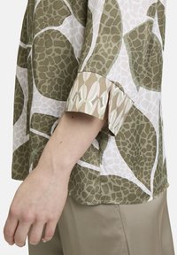 Groene en witte gebloemde blouse met korte mouwen, versierd met een loofmotief en een zijsplit aan de onderkant. Zachte stof en een relaxte pasvorm.
