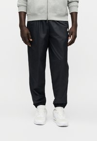 Μαύρο, ελαφρύ αθλητικό παντελόνι με σούρες στα τελειώματα, με μικρό λογότυπο στην αριστερή πλευρά. Φορεμένο με λευκά αθλητικά παπούτσια και γκρι μπουφάν με φερμουάρ.