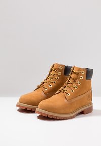 Bottes en suede beige avec un design à lacets, œillets dorés, col rembourré et semelle en caoutchouc avec un relief texturé. Logo Timberland sur le côté.
